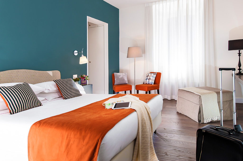 Quarto na Corso Vittorio 308 com cama de casal, janela, duas cadeiras coloridas, televisão, mesa de cabeceira e abajur.